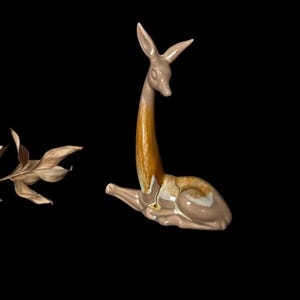May include: A ceramic deer figurine, beige with brown and gold accents, in a seated pose. The deer has a long neck and slender legs. A dried leaf branch is to the left of the deer against a black background.