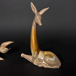 Peut inclure: Figurine de cerf en céramique au repos, avec un long cou et des oreilles pointues. Le cerf a un corps beige avec un cou marron et doré. Une branche de feuilles séchées est en arrière-plan. La figurine est sur une surface noire.