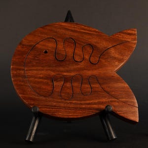 May include: A wooden fish-shaped puzzle with a dark brown finish, displayed on a black metal stand. The puzzle has a carved design within its body. A few pink flower petals are on the black surface.