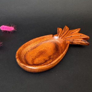 May include: A wooden pineapple-shaped bowl, hand-carved with an oval body and a pineapple top. The wood has a warm, reddish-brown colour. The bowl sits on a dark background with pink decorative accents.