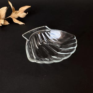 May include: A clear, seashell-shaped glass dish with a ridged design. The dish sits on a dark surface, with a dried plant stem in the upper left corner. The dish is likely for serving or display.