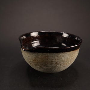 May include: A small, brown and white ceramic bowl with a slightly uneven rim. The bowl has a dark brown glaze on the inside and a lighter brown glaze on the outside.