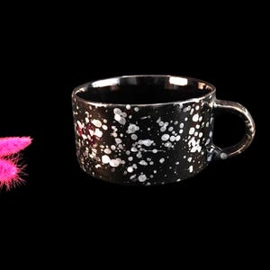 May include: A black ceramic mug with white speckled paint. The mug has a rounded shape with a handle. Two bright pink, fluffy decorative stems are to the left of the mug against a black background.