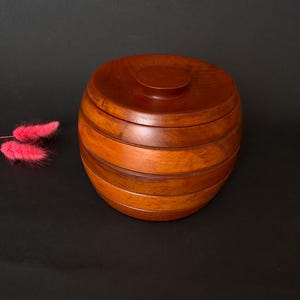 Peut inclure: Une boîte ronde en bois brun chaud avec un couvercle. La boîte est composée de bandes horizontales de bois, créant une texture. Un petit accent floral rose est visible en arrière-plan.