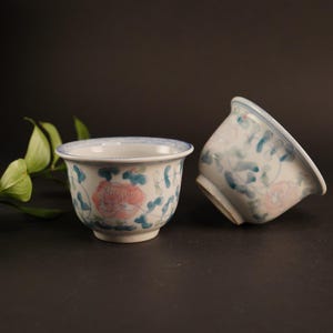 May include: Two white ceramic teacups with blue and pink floral designs. The cups are sitting on a black surface with a green leafy branch.