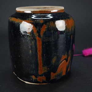 May include: A dark blue ceramic jar with brown and white glaze accents. The jar has a rounded rectangular shape and a wide opening.