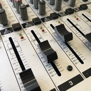 May include: Close-up of a silver mixing console with multiple faders and knobs. The faders are labelled with numbers 2, 3, 4, 5/6, 7/8, and the knobs are labelled with the words "PAN", "AUX", "MUTE", "ALT 3-4", and "PEAK".