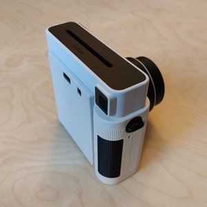 May include: A white Polaroid Now+ instant camera with a black lens and a black film pack slot. The camera has a black and white striped design on the side.