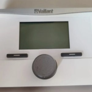 May include: White thermostat with a digital display and a round knob. The thermostat has the word "Vaillant" printed on it.
