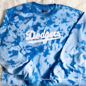 May include: A long-sleeved, tie-dye shirt in shades of blue. The word "Dodgers" is printed across the chest in white, stylized lettering. The shirt has a crew neck and long sleeves. The overall design is a blend of light and dark blue hues.