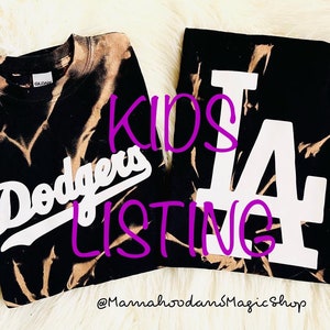 May include: Two black t-shirts with bleach-dyed patterns. One shirt features the word "Dodgers" in white script, and the other displays a large white "LA" logo. The text "KIDS LISTING" is in purple. The shop's name is also visible.