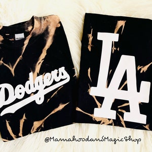May include: Two black sweatshirts with bleach-dye patterns. One features the word "Dodgers" in white script, and the other displays a white "LA" logo. The sweatshirts are on a white, fluffy surface.