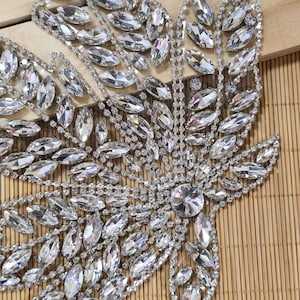 May include: A large, leaf-shaped appliqué featuring a dense arrangement of clear, marquise-cut rhinestones set in silver-toned metal. The design includes a central circular stone with radiating rows of crystals forming a detailed, symmetrical pattern.