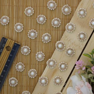 May include: A close-up of a collection of pearl and gold embellishments. The embellishments are arranged on a wooden surface and a ruler is visible in the foreground.