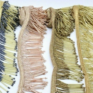 May include: A selection of beaded fringe trim in gold, black, and rose gold shades. The trim, with its beaded strands, is suitable for embellishing clothing, accessories, or home decor. The fringe is displayed on a white surface. Measurements are in centimetres.