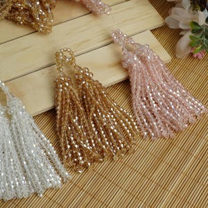 May include: Close-up of beaded tassels in white, gold, and pink hues. The tassels are made of small, faceted beads strung together, creating a shimmering effect. They are arranged on a light brown wooden surface and a bamboo mat.