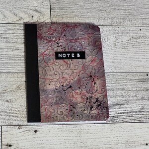 May include: A notebook with a textured cover featuring a floral design in shades of gray, pink, and purple. The word "NOTES" is displayed on a black rectangle. Black ink splatters add detail to the cover.