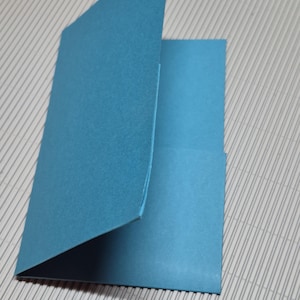 May include: A partially opened, teal-colored card. The card is made of thick paper and has a clean, simple design. The card is on a light gray surface with parallel lines.