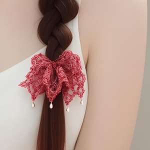 Puede incluir: Un accesorio para el cabello con forma de lazo de encaje rojo con dijes colgantes en forma de lágrima está sujeto a una trenza marrón oscuro. El lazo está hecho de un material delicado de tejido abierto.