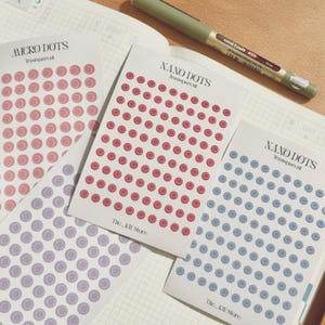 May include: Three rectangular sheets of transparent adhesive dot stickers in pink, red, and blue, each labelled Nano Dots or Micro Dots with The KW Store printed at the bottom. A green Uni-ball eye fine pen rests on a grid-patterned notebook page.