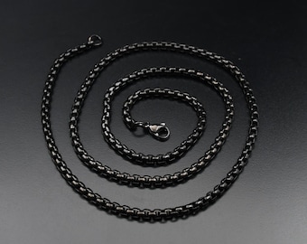 May include: A black box chain necklace with a lobster clasp. The necklace is made of small, interconnected links and is displayed on a dark background. The chain is a dark, matte black colour.