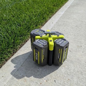 May include: A multi-battery power tool charger with four black and yellow 40V lithium batteries. The charger has a yellow handle and is sitting on a concrete surface next to green grass.