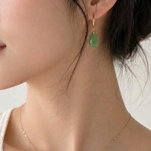 May include: A gold drop earring featuring a smooth, teardrop-shaped green stone suspended from a thin gold wire hook. The earring is worn on the earlobe, and a delicate gold chain necklace with small, flat, reflective links rests on the neck.