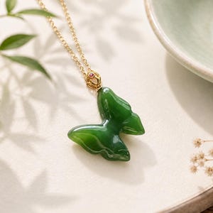 May include: A necklace featuring a carved green jade butterfly pendant suspended from a gold chain. The pendant is attached to the chain by a gold bail accented with a small, round, deep pink gemstone at the top of the butterfly wings.
