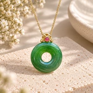 May include: A gold chain necklace featuring a circular green jade pendant. The pendant is topped with a gold decorative bail accented by a small, round, pink gemstone. The necklace is suspended from a thin gold link chain with a polished finish.
