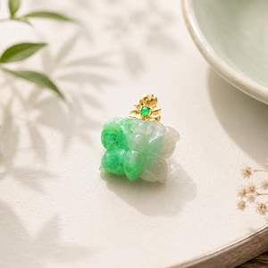 May include: A butterfly-shaped pendant carved from translucent green and white jadeite. The piece features a small gold-toned metal accent at the top, adorned with a tiny, round, vibrant green stone set in the center of a floral-shaped gold element.