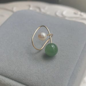 Könnte beinhalten: Ein goldfarbener Ohrklemme mit einer weißen Perle und einer grünen Jadekugel. Das Design hat eine zarte, offene Form. Der Schmuck wird auf einer grauen Oberfläche präsentiert.