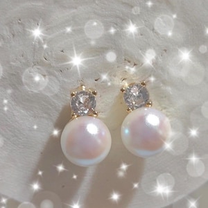 Puede incluir: Un par de pendientes de oro con una perla y un cristal transparente. Los pendientes están sobre un fondo blanco.