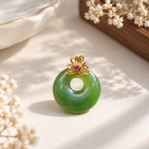 May include: A circular green jade pendant featuring a polished donut shape. The pendant is topped with a gold filigree bail in a floral design, accented by a small, round, vibrant pink gemstone set at the center of the gold metal structure.