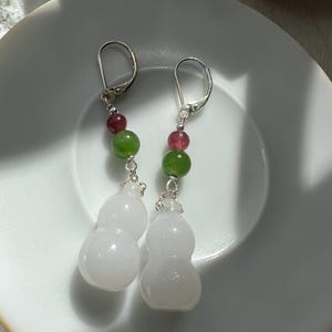 May include: A pair of dangle earrings featuring white gourd-shaped pendants. Each earring has a silver hook, with a strand of green and red beads. The earrings are displayed on a white plate.
