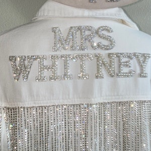 May include: White denim jacket with a rhinestone fringe trim. The jacket has a large rhinestone appliqué that says "MRS WHITNEY". A white cowboy hat with a rhinestone appliqué that says "BRIDE" is on top of the jacket.