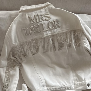 Puede incluir: Chaqueta vaquera blanca con el texto "MRS TAYLOR" en adornos perlados en la espalda. La chaqueta presenta flecos plateados en la espalda y en las mangas, creando un look glamuroso.