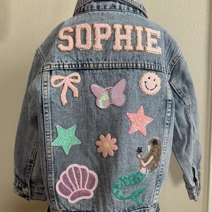 Op de afbeelding: Een lichtblauw denim jack met de naam "SOPHIE" in roze glitterletters op de rug. Het jack is versierd met verschillende kleurrijke patches, waaronder een vlinder, een ster, een schelp, een bloem, een zeemeermin, een strik, een smiley en een ster.