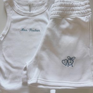 Może przedstawiać: Biały top na ramiączkach i pasujący zestaw szortów. Top ma wyhaftowany napis "Mrs. Fischer" w jasnoniebieskim kolorze. Szorty mają dwie inicjały w kształcie serca ze wstążką.