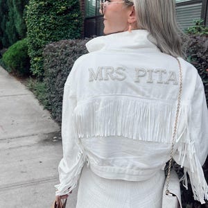 以下が含まれることがあります： フリンジ付きの白いデニムジャケットと、パールで装飾された「MRS PITA」の文字。ジャケットは、お揃いの白いショートパンツと組み合わせています。ゴールドチェーンの白いハンドバッグが見えます。