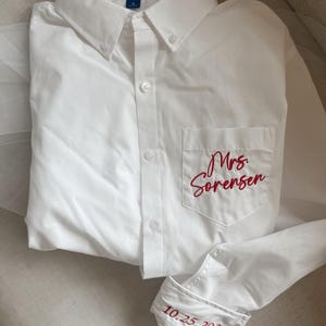 Pode incluir: Uma camisa branca abotoada com a marca "Port Authority" na gola. A camisa tem um bolso com o nome "Mrs. Sorensen" bordado em vermelho. A manga mostra a data "10.25.2025" em vermelho.
