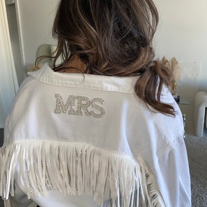 May include: White denim jacket with fringe trim and a rhinestone 'MRS' embellishment on the back.