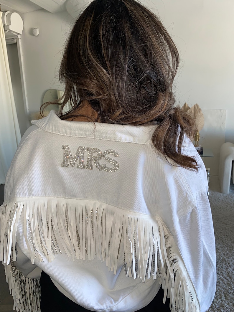 May include: White denim jacket with fringe trim and a rhinestone 'MRS' embellishment on the back.