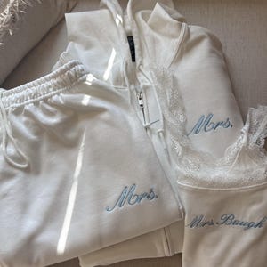 May include: A white zip-up hoodie, white sweatpants with a drawstring, and a white ribbed camisole with lace trim. The hoodie and sweatpants feature the embroidered blue text "Mrs." while the camisole features the embroidered blue text "Mrs. Baugh."