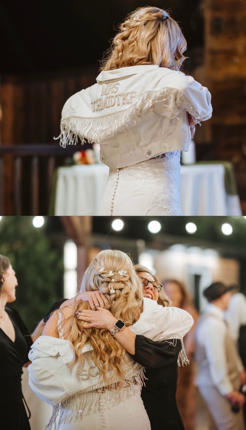 May include: A bride wearing a white wedding dress and a white denim jacket with "Mrs. Schmidtke" embroidered on the back. The bride is hugging another person wearing a black dress.
