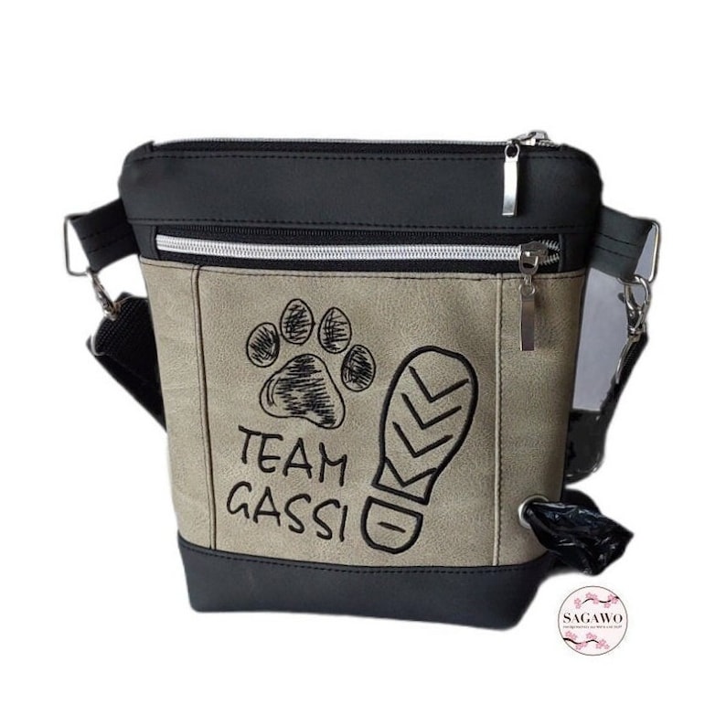 May include: Black and beige crossbody bag with a zipper closure. The bag has a paw print and a footprint embroidered on it with the text "Team Gassi".