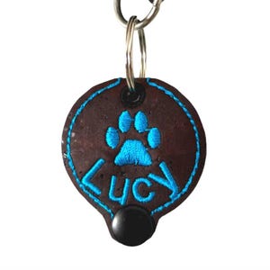 Könnte beinhalten: Ein braunes Leder-Hundehalsband mit einem blauen Pfotenabdruck und dem Namen "Lucy" bestickt. Das Halsband ist an einem silbernen Schlüsselring befestigt.