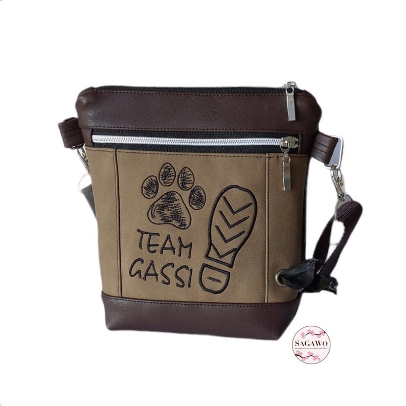 May include: A brown and tan canvas crossbody bag with a zipper closure. The bag has a black embroidered paw print and the text "TEAM GASSI" on the front. The bag has a brown leather trim and a black adjustable strap.