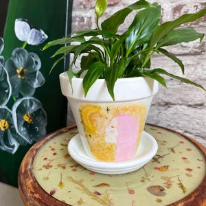 Może przedstawiać: Biała ceramiczna doniczka z pasującą podstawką, zawierająca zieloną roślinę skrzydłokwiat. Doniczka ozdobiona jest ręcznie malowaną ilustracją profilu z różowymi włosami na żółtym, fakturowanym tle, z motywem półksiężyca i gwiazd.