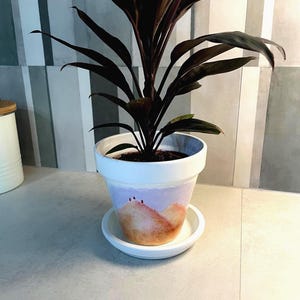 Puede incluir: Una maceta de cerámica blanca con su plato a juego, decorada con un paisaje pintado a mano de colinas naranjas y amarillas bajo un cielo morado claro. Una planta de interior con hojas oscuras y puntiagudas crece desde el centro del tiesto.