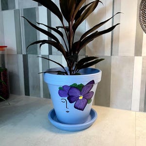 Op de afbeelding: Een lichtblauwe keramische plantenpot met bijpassende schotel, voorzien van een handgeschilderd ontwerp van een paarse viooltjesbloem boven een minimalistische zwarte lijntekening van een menselijk gezichtsprofiel, met een kamerplant.
